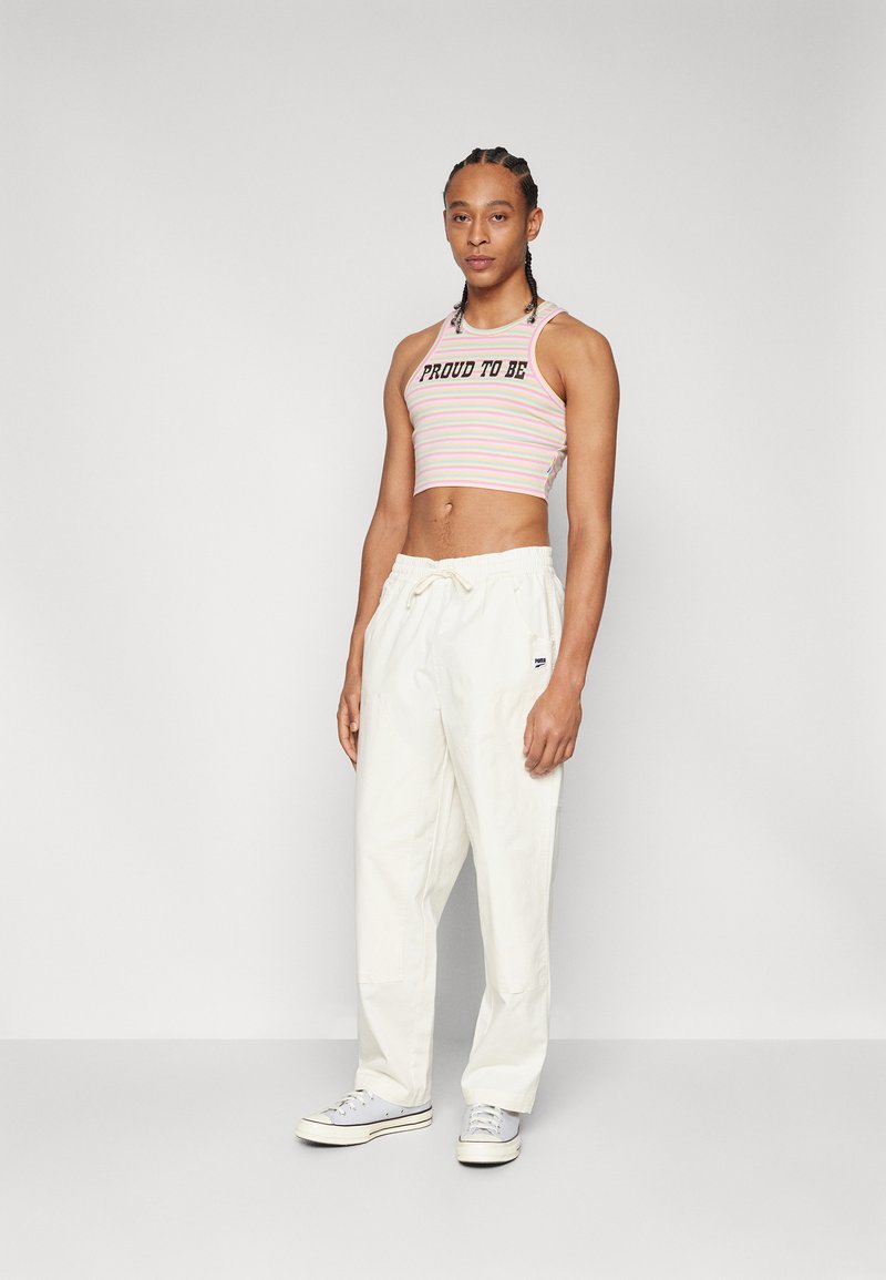 Cropped, multicolored striped tank top with text "PROUD TO BE" paired with loose-fitting white trousers and gray sneakers.