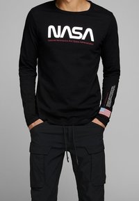Camiseta de manga larga negra con el logo en blanco de "NASA" y texto en rojo. Presenta un parche de la bandera americana en la manga. Material de algodón, diseño ajustado.