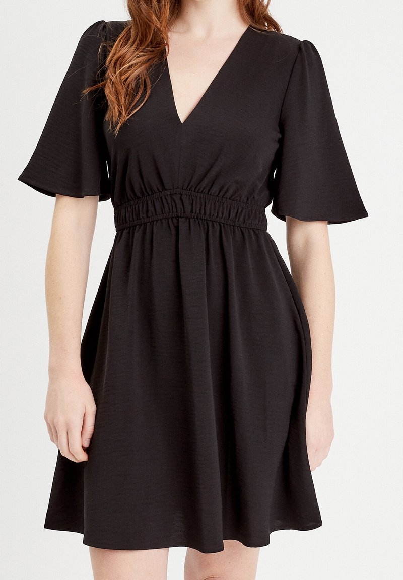 Femme portant une robe noire à manches courtes avec un décolleté en V et une taille élastiquée, montrée des épaules jusqu'à mi-cuisse.