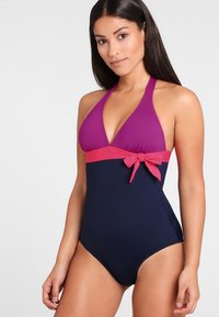 Eéndelige zwemoutfit met een paarse halternek en een roze band met een strikje rond de taille, tegen een marineblauwe ondergrond. Soepele stoftextuur.