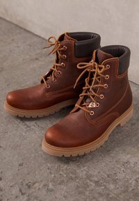 Botas de cuero marrón con cordones, cuello acolchado, suela de goma beige y detalles cosidos. Presentan cordones redondos y ojales de metal.