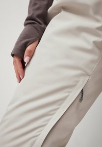 Beige broek met een gladde textuur en een zijrits. Een hand van een persoon rust op het been. De mouw is bruin, nauwsluitend, met een duimgat.