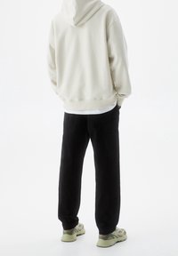 Persona che indossa un hoodie beige, pantaloni cargo neri e sneakers chiare, in piedi con le mani in tasca, rivolto di spalle alla macchina fotografica.