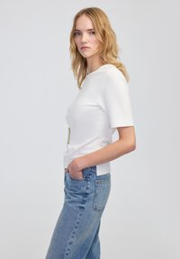 Weißes Kurzarm-Oberteil mit einem runden Halsausschnitt, ausgestattet mit einem dekorativen Akzent aus goldfarbenem Metall und einem lässigen Schnitt; kombiniert mit hellblauen Jeans.