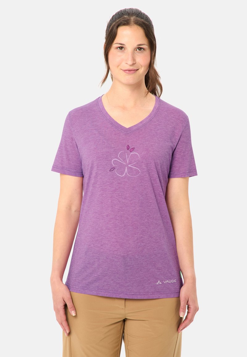 Donna con capelli castani raccolti in una coda di cavallo, che indossa una maglietta viola a V con un motivo floreale e pantaloni beige, in piedi davanti a uno sfondo semplice.