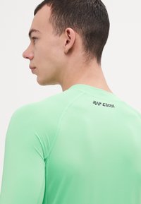 Ilgi piedurkņu, gaiši zaļš saules aizsargapģērbs, izgatavots no elastīga auduma. Tam ir plakans šuvums un mazs "RIP CURL" logo aizmugurējā kakla daļā.