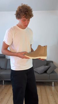 Bež sametist ankle boot elastsete külgpaneelidega, valge kummitald ja sile pind, hoiab isik valges T-särgis.