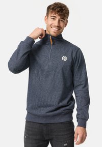 Marineblå trøje med halvlukke krave, stribet tekstur, logo på brystet og ribbede manchetter og kant.