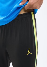 Käsi tõmbab üles sinise särgi, rõhutades musti sportpükse kollase korvpallimängija logoga ning kollase küljeribaga.