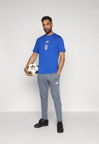 Μπλε μπλούζα Adidas με το σήμα της Ιταλίας και χρυσές λεπτομέρειες, σε συνδυασμό με γκρι φόρμες Adidas με πλευρικές λωρίδες, κρατώντας μια μπάλα ποδοσφαίρου.