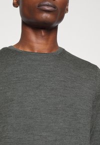 Maglione a maglia grigio scuro con scollatura rotonda e texture sottile. Presenta un piccolo logo sulla parte anteriore, aderente al corpo.