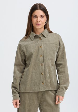 Femme aux cheveux bruns lisses portant une chemise boutonnée en velours côtelé vert olive et un pantalon assorti, debout devant un fond uni clair.