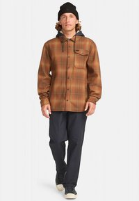 Bruine geruite flanellen overhemd met een contrasterende marineblauwe capuchon, knoopsluiting en borstzak. Gestyled met donkere broek en casual schoenen.