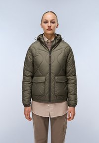 Olijfgroene gewatteerde pufferjack met capuchon, voorzien van een rits aan de voorkant, twee grote zakken en een aansluitende zoom. Draagt over een beige shirt.