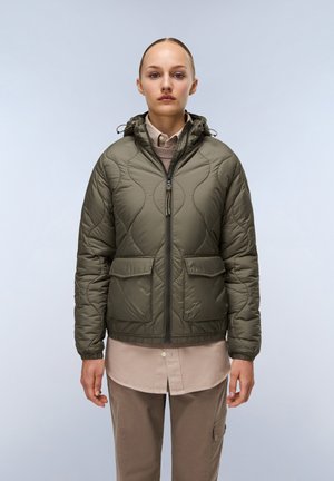 Olijfgroene gewatteerde pufferjack met capuchon, voorzien van een rits aan de voorkant, twee grote zakken en een aansluitende zoom. Draagt over een beige shirt.