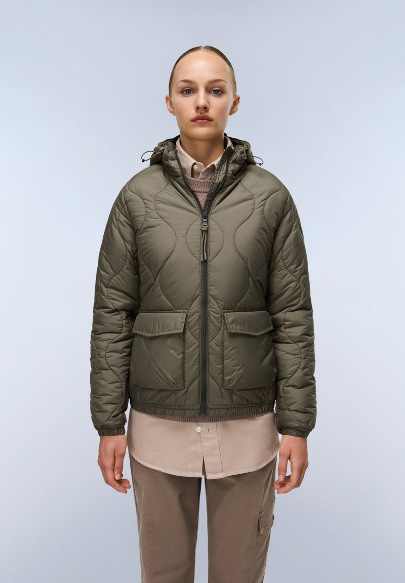 Olijfgroene gewatteerde pufferjack met capuchon, voorzien van een rits aan de voorkant, twee grote zakken en een aansluitende zoom. Draagt over een beige shirt.