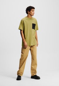 Alie žalios spalvos marškiniai su juodu krūtinės kišeniu, derinami su smėlio spalvos laisvais džinsais ir juodais batais, stovintys ant vientiso balto fono.