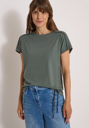 Jeune femme aux cheveux bruns portant un haut vert à manches courtes avec des liens sur les côtés et un jean bleu, se tenant devant un fond clair uni.