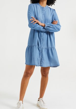 Kobieta ze skrzyżowanymi ramionami, ubrana w jasnoniebieską, zapinaną na guziki sukienkę jeansową i białe trampki, stojąca na tle jednolitego, białego tła.