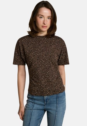 Vrouw met schouderlang bruin haar, gekleed in een bruin shirt met korte mouwen en luipaardprint en een blauwe spijkerbroek tegen een effen achtergrond.