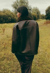 Pruun nahast jakk laia kraega ja pehme, tekstureeritud siseosaga, kantud üle siledast tumedast kangast kleidi, välirohelises rohumaas.