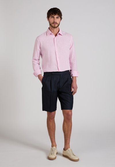 Man wearing light pink long-sleeve button-up shirt, black pleated shorts, and beige sneakers standing with hands in pockets.
