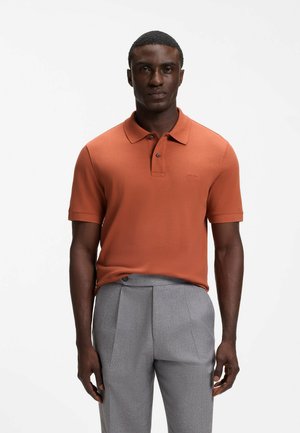 Mann trägt ein kurzärmeliges Polo-Shirt in verbranntem Orange und hellgraue plissierte Hose vor weißem Hintergrund.