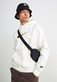 Hanorac alb cu glugă, având un logo Nike în nuanțe de galben și negru, asortat cu o pălărie tip bucket neagră și o geantă crossbody neagră. Pantaloni maro purtați în partea de jos.