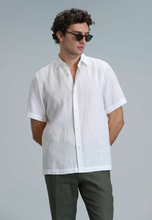 Chemise en lin blanche à manches courtes avec un col boutonné, texture lisse et légères rides. Portée avec un pantalon vert foncé et des lunettes de soleil.