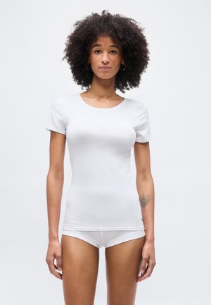 Γυναίκα με σγουρά μαλλιά, φορώντας στενό, λευκό πουκάμισο με κοντά μανίκια και λευκά εσώρουχα, που στέκεται μπροστά σε απλό ανοιχτό φόντο.