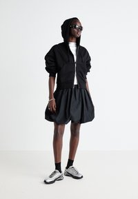 Felpa nera con zip e cappuccio, abbinata a una gonna nera voluminosa. Completata da scarpe sportive argento e calze scure.