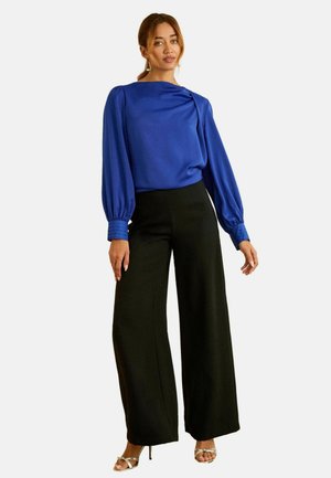 Blusa de satén azul royal con mangas abullonadas y escote redondeado, combinada con pantalones de pierna ancha negros de cintura alta y tacones metálicos.
