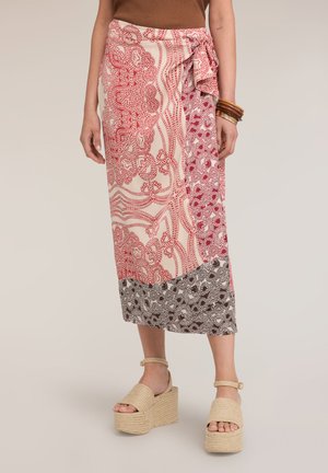 Kvinde iført et rødt og pink mønstret omlægning-nederdel med en sløjfe i siden, beige platformsandaler og stablede træarmbånd på det ene håndled.