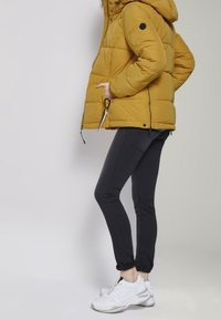 Hořčicově žlutá péřová bunda s kapucí a zipovými kapsami, kombinovaná s černými úzkými kalhotami a bílými teniskami. Přiléhavý střih, prošívaná textura.
