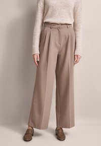 Pleated wide-leg trousers in light brown fabric, featuring a high waist and tailored fit. Accompanied by a beige sweater and brown loafers.