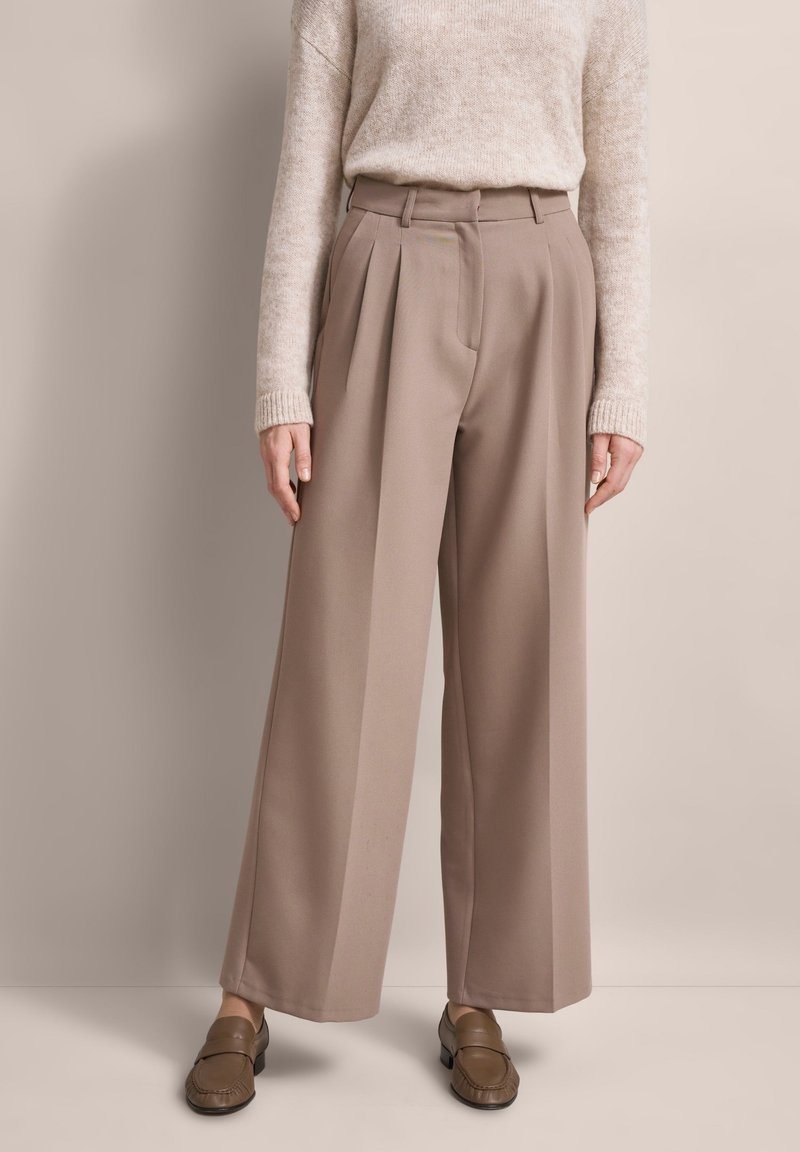 Pleated wide-leg trousers in light brown fabric, featuring a high waist and tailored fit. Accompanied by a beige sweater and brown loafers.