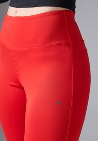 Lyse røde leggings laget av elastisk stoff med høy midje, flatlock-sømmer og en liten, mørk logo-detalj på låret.