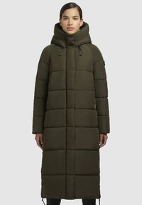 Ženska nosi dolgo olivno zeleno podlo�ženo zimsko jakno z visokim ovratnikom in vrvico na kapuci, stoji pred enostavno svetlo ozadje.