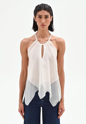 Femme aux cheveux courts et foncés porte un haut blanc sans manches à col halter avec des panneaux transparents superposés et un pantalon bleu marine sur fond uni.