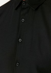 Zwarte gekraagde shirt, textuurstof, met vijf zwarte knopen en een knoopsluiting. Close-up van het shirtdetail.