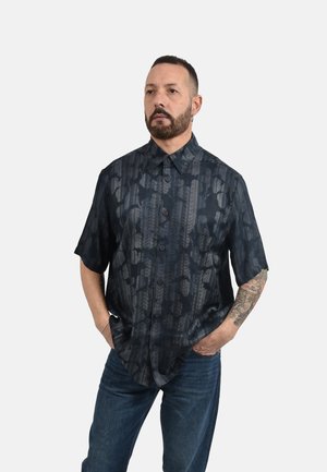 Mężczyzna z brodą i tatuażem na ramieniu, ubrany w ciemną koszulę z krótkim rękawem z wzorem oraz niebieskie dżinsy, ręce w kieszeniach, spogląda lekko na bok.