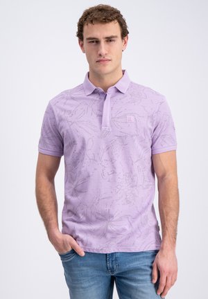 Giovane uomo che indossa una polo fantasia viola chiaro con colletto e jeans blu, in piedi con una mano in tasca su sfondo bianco.