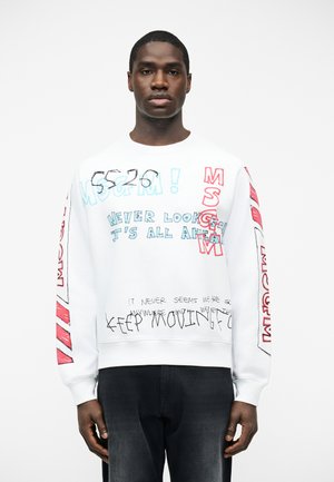 Homme noir portant un sweat-shirt blanc avec du texte manuscrit multicolore et des motifs graphiques rouges, debout devant un fond blanc uni.