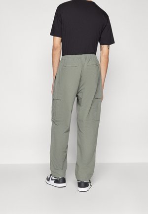 Hombre vistiendo pantalones cargo verde oliva con bolsillos traseros y zapatillas blancas y negras, de pie contra un fondo claro y liso.