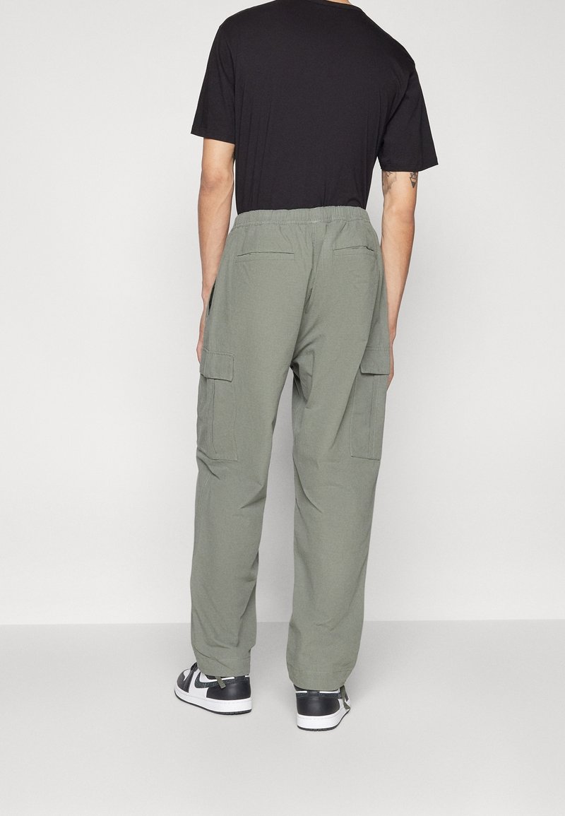 Homme portant un pantalon cargo vert olive avec des poches arrière et des baskets noires et blanches, debout devant un fond clair uni.