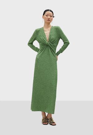 Long green dress made of textured fabric, featuring a deep V-neck and a knot detail at the waist. Model paired with embellished black sandals.