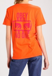 Camiseta de algodón naranja de manga corta que presenta un gran gráfico rosa en la espalda que dice "NO OBEDEZCAS A NADIE" con símbolos de mano.