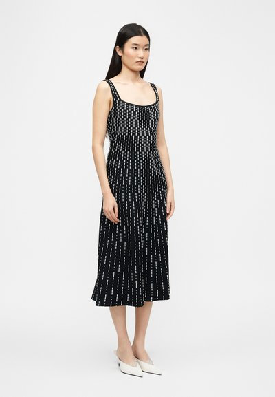 Γυναίκα με μακριά σκούρα μαλλιά, που φορεί μαύρο αμάνικο midi φόρεμα με λευκές κάθετες τελείες και λευκά παπούτσια με μυτερή μύτη, στέκεται μπροστά σε λευκό φόντο.