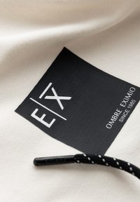 Étiquette rectangulaire en tissu noir avec logo blanc "EX" et texte "OMBRE EXIMIO SINCE 1985" sur tissu clair, cordon noir à pois blancs à proximité.