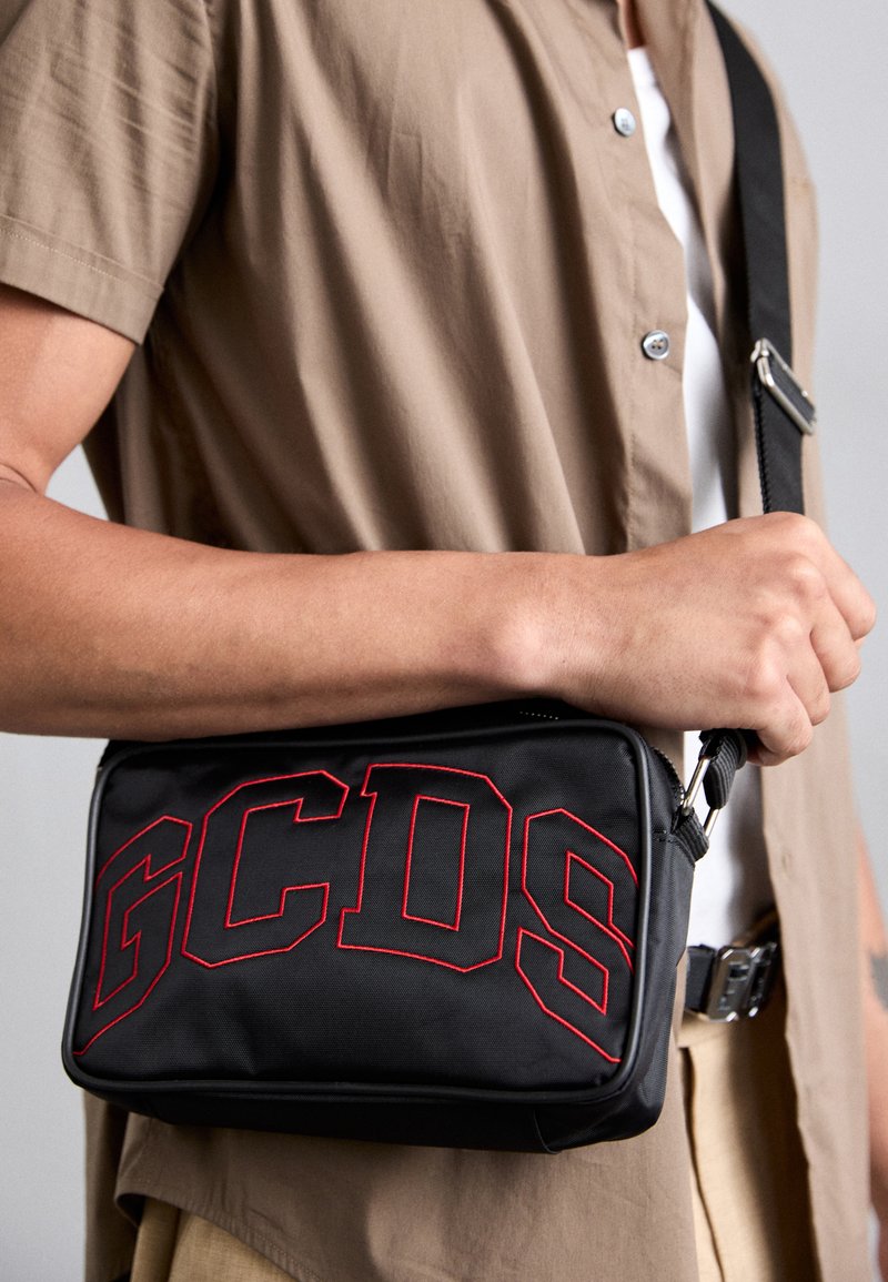 Črna tkaninska torba čez ramo z rdečim vezenim logotipom "GCDS" spredaj, nastavljivim trakom in zadrgo. Gladka tekstura.